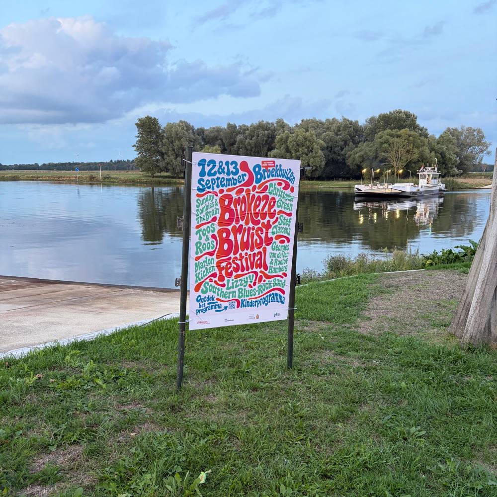 Brokeze Bruist Festival poster mounted between two metal posts on a grassy riverbank at dusk, with a calm river and a small ferry in the background. The poster’s melting, retro lettering lists 12 and 13 September in Broekhuizen and the performers, with a QR code at the bottom.