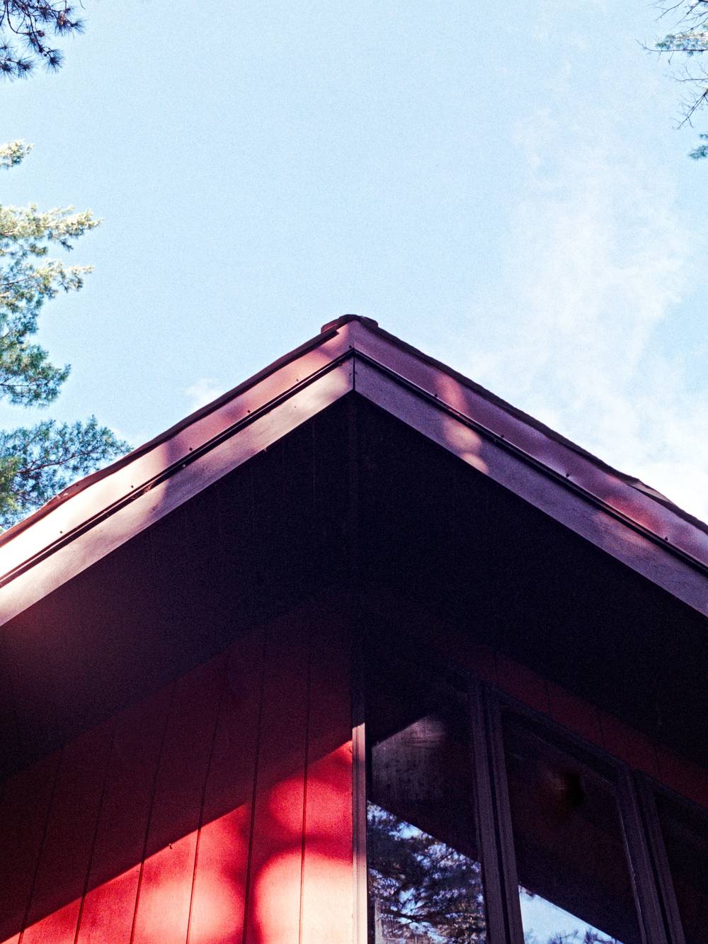 The sharp corner of a red building roof points upward against a clear blue sky, framed by the silhouettes of pine trees.