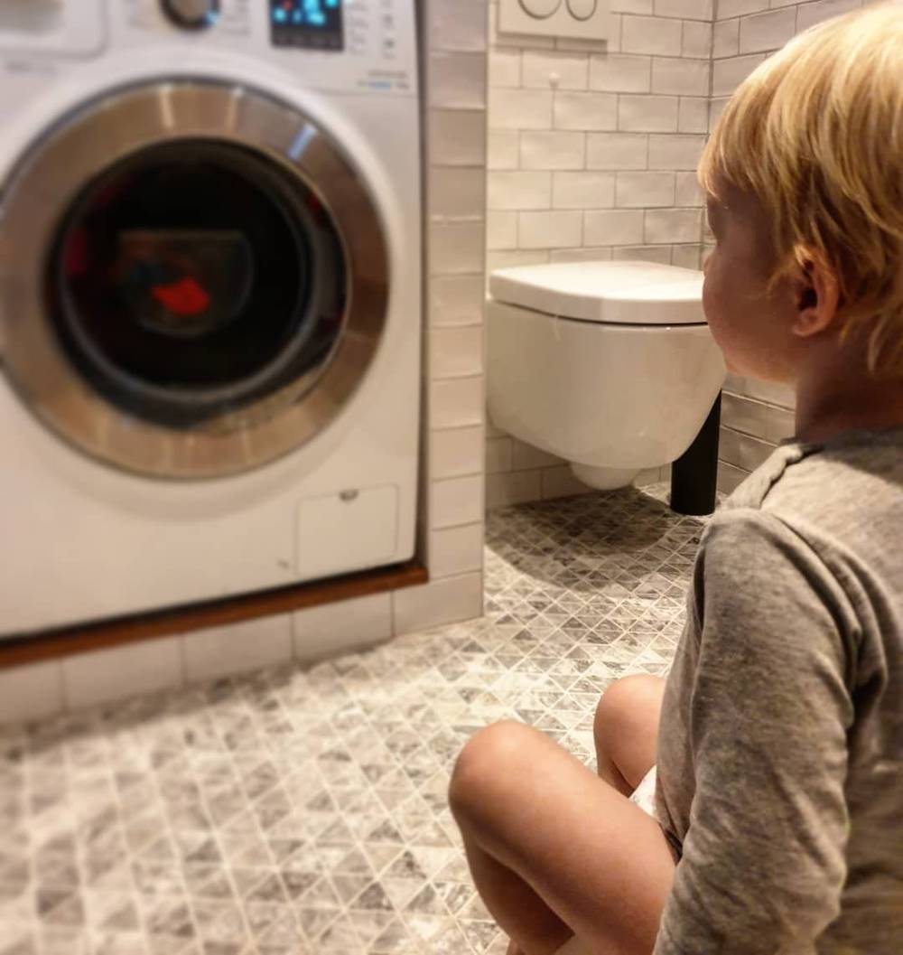 A child sitting on a tiled bathroom floor looking up at a wall-mounted toilet.