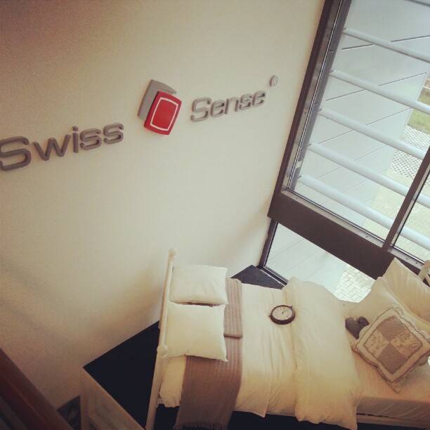 A hotel room interior with a made bed and pillows next to a 'Swiss Sense' branded wall sign.