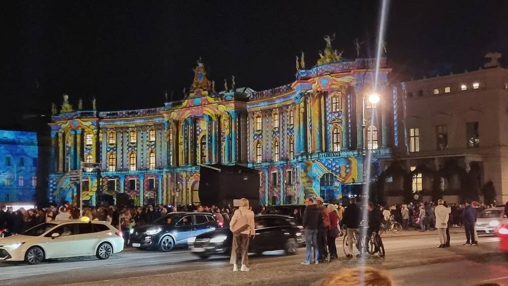An ornate building facade lit up with colorful projected patterns at night, with a crowd and cars gathered in front.