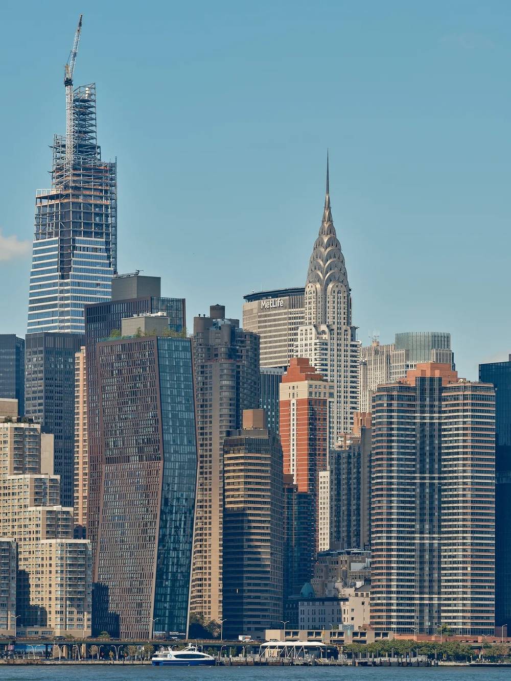 The Midtown Manhattan skyline viewed from across the East River under a clear blue sky. One Vanderbilt tower is visible under construction on the upper left, its steel frame topped with a construction crane. The iconic Art Deco Chrysler Building with its distinctive stainless steel eagle crown and spire rises prominently in the center of the skyline. The MetLife Building is visible behind it. A cluster of modern glass and brick residential and commercial towers fills the dense skyline. A ferry and waterfront promenade are visible along the river's edge in the foreground.