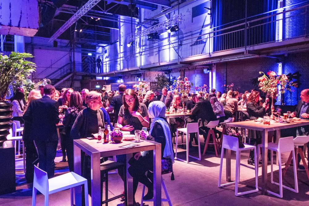 A crowd gathers around tables with floral centrepieces in the blue-lit hall at De Fabrique.