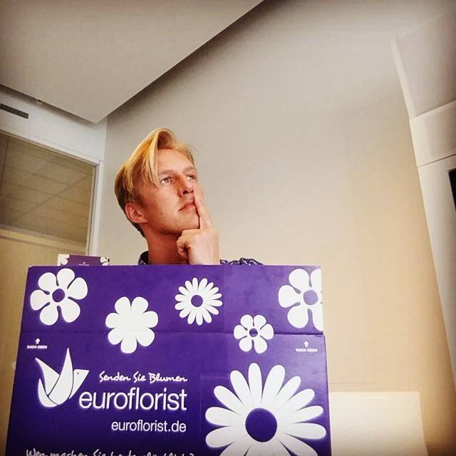 A man rests his chin on his hand, seated behind a purple gift bag branded 'Euroflorist'.