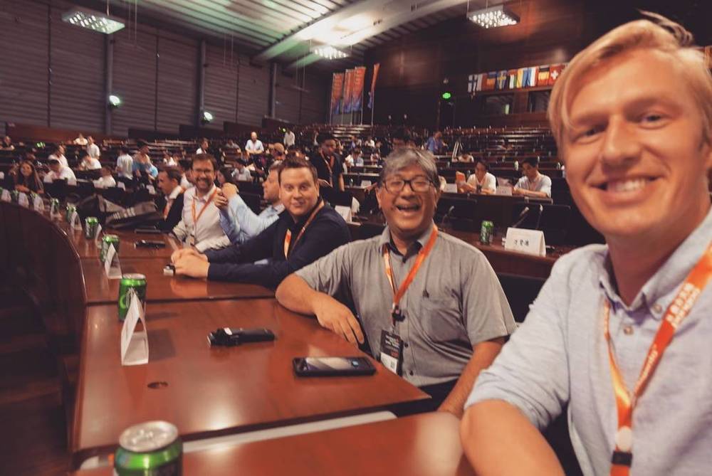 A group of men posing at a conference table with drinks, in a crowded auditorium.