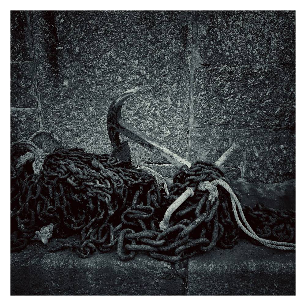 A black and white photograph of an old, weathered anchor resting against a rough stone wall, surrounded by tangled piles of heavy iron chains and thick rope.