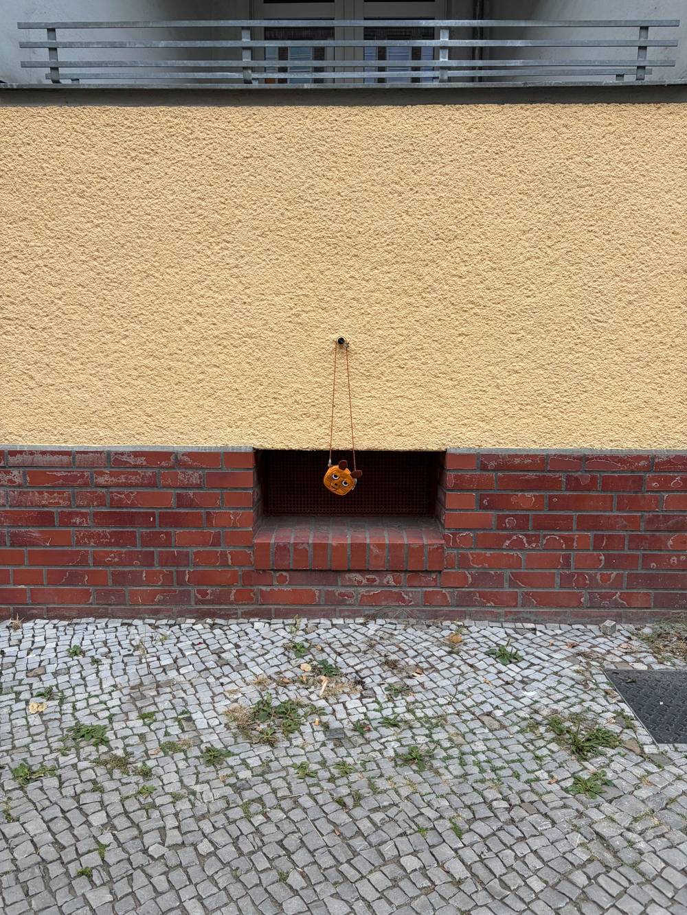 ein kinderportemonaie hängt an einem band vor einem kellerfenster. das foto wird dominiert von horizontalen linien der fassade, der gelbe putz fällt als erstes in auge, drunter ein rotes band ziegelsteine, weiter unten graues Pflaster.
