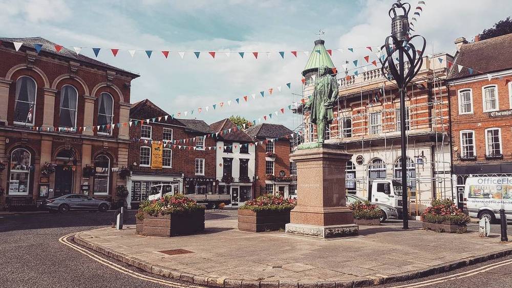 A market square in an English town decorated with colorful bunting, flower planters and a statue.