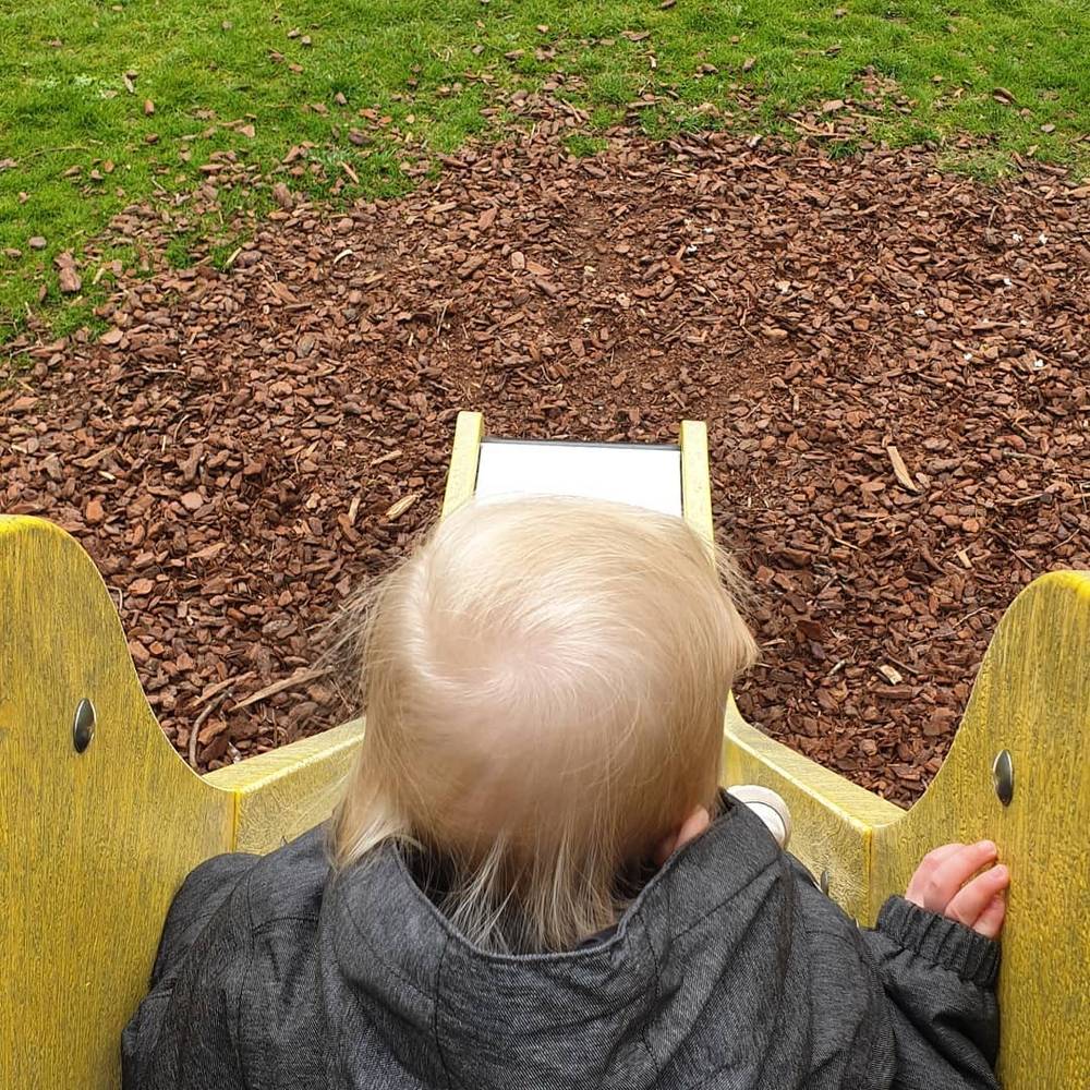 Looking down at a toddler's blond head at the top of a yellow playground slide, wood chips scattered below.