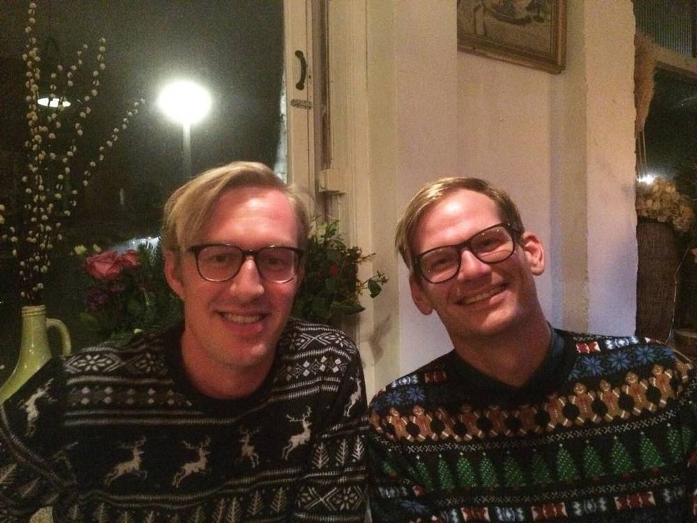 Two men in Christmas sweaters posing together indoors at night.