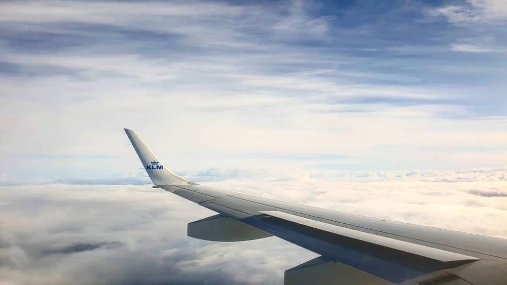 View from an airplane window above the clouds, a KLM logo visible on the wing.