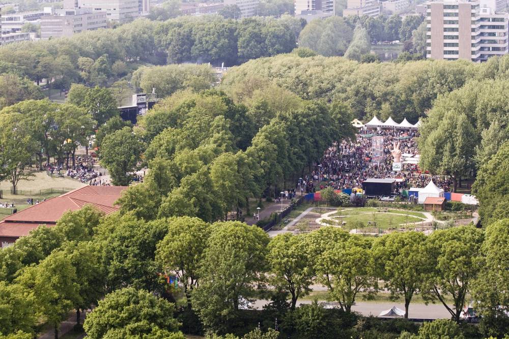 An aerial view of a tree-lined park with a distant festival stage and crowd.