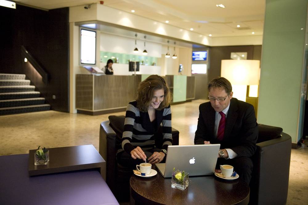 A modern hotel lobby with a reception desk, staircase and two people seated with laptops.