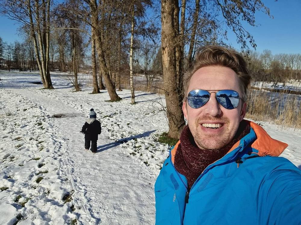 Guido smiling in a blue jacket, with a bundled-up child walking through a snowy park behind him.