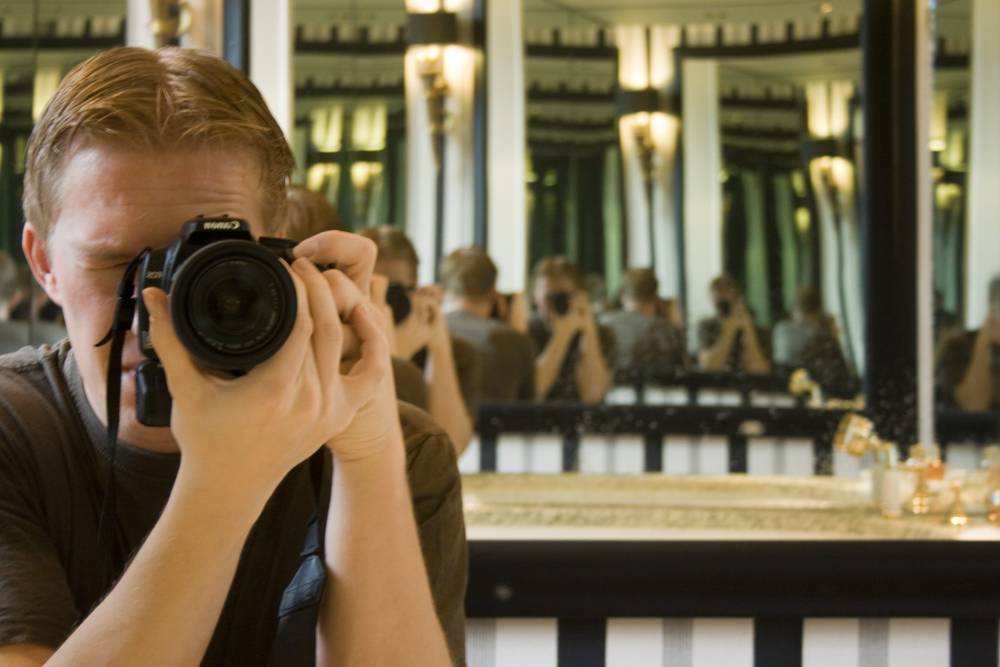 A man photographing himself in a mirror, with a reflected group of other photographers behind him.