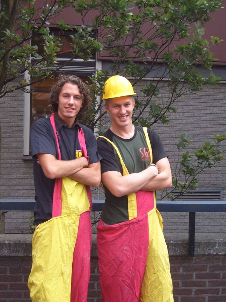 Two young men pose in colorful rubber overalls and a hard hat outdoors.