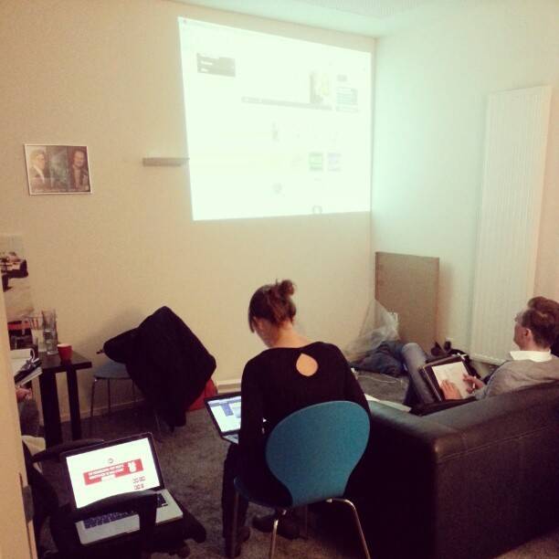 Two people work on laptops on a sofa while a presentation is projected on the wall behind them.