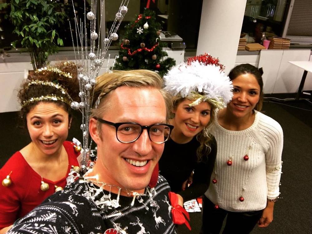 A selfie of a smiling man wearing festive tinsel and Christmas lights with three colleagues in holiday headbands.