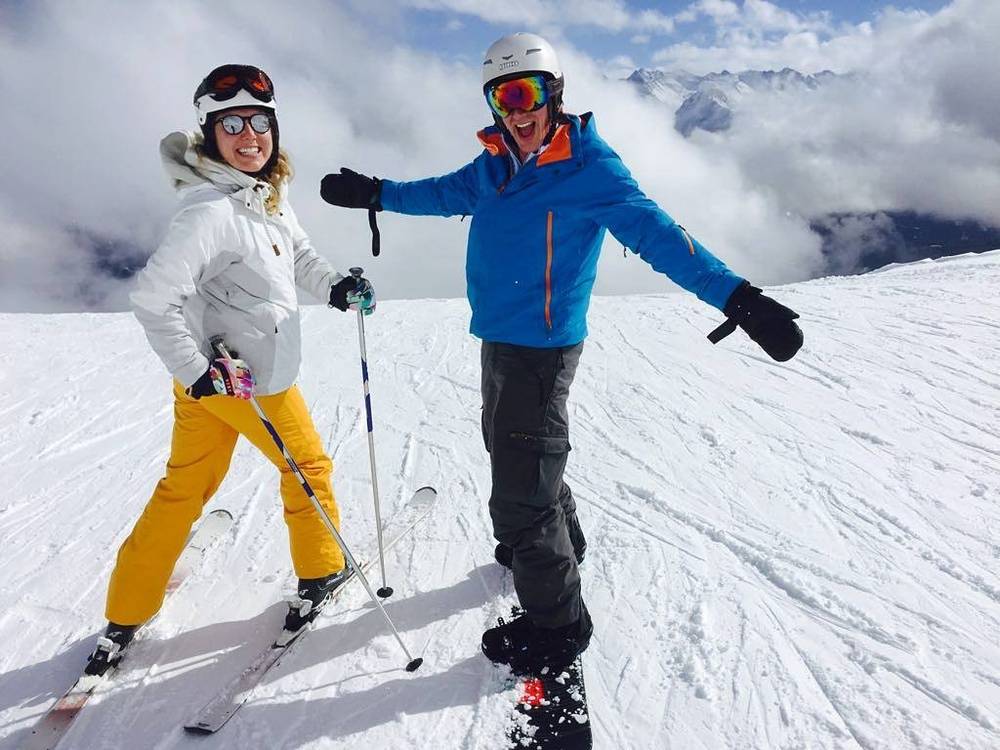 Two skiers pose together on a snowy slope with mountains behind them.