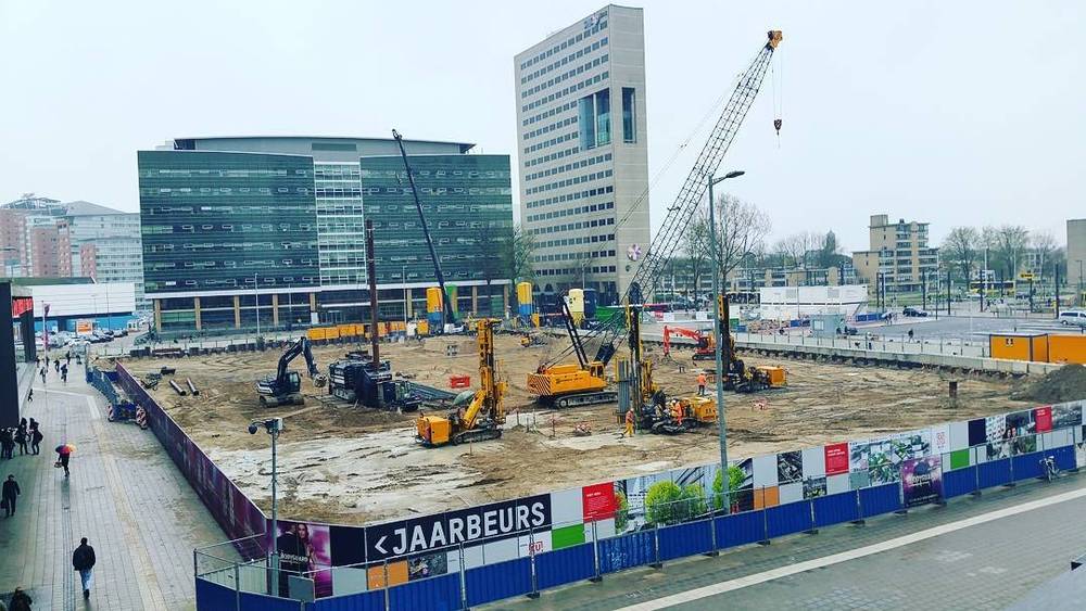 Construction site with cranes and diggers next to office towers, cordoned off by a Jaarbeurs fence.