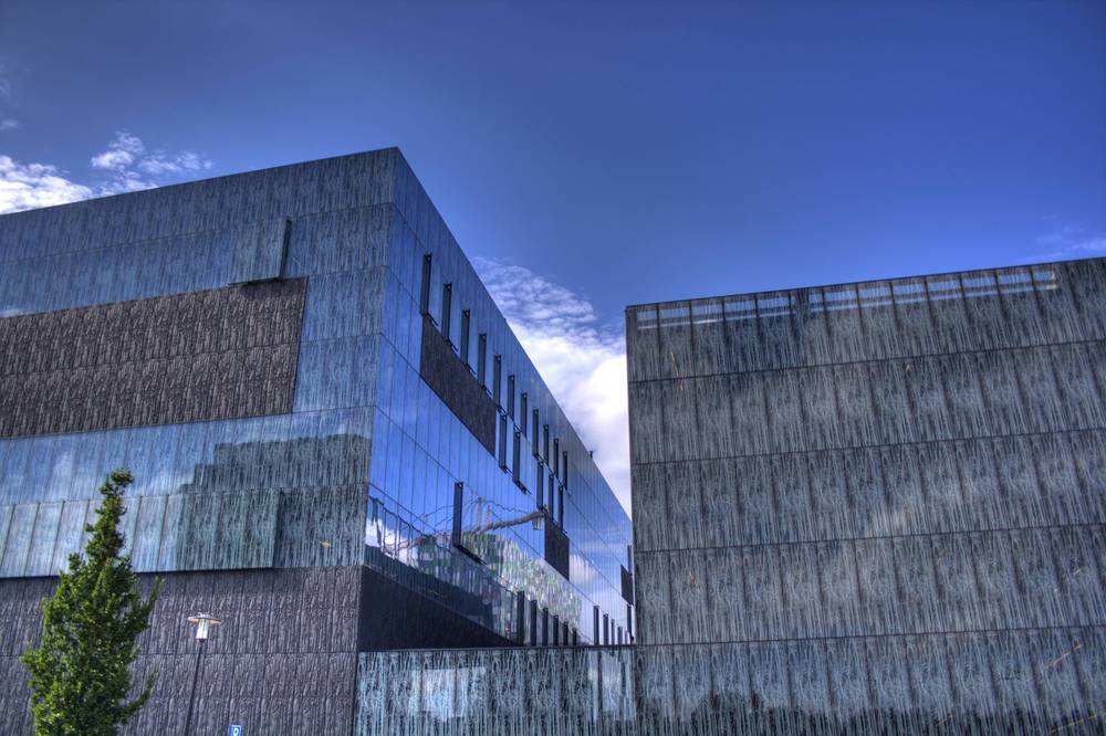 Angular glass and metal-clad university buildings meeting at a sharp corner under a blue sky.