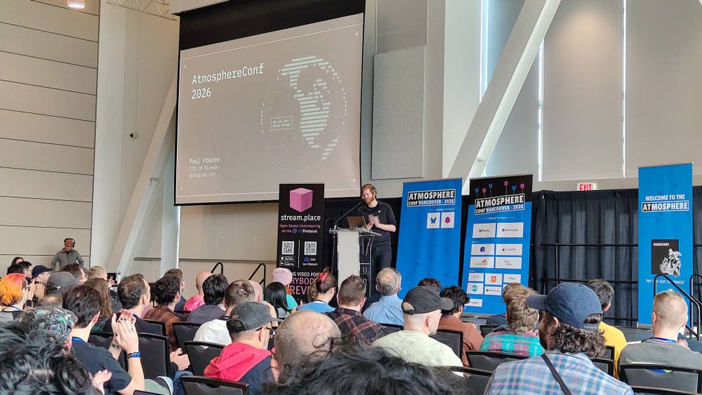 An audience listens to a speaker at a podium, with a screen reading "AtmosphereConf 2026" behind.