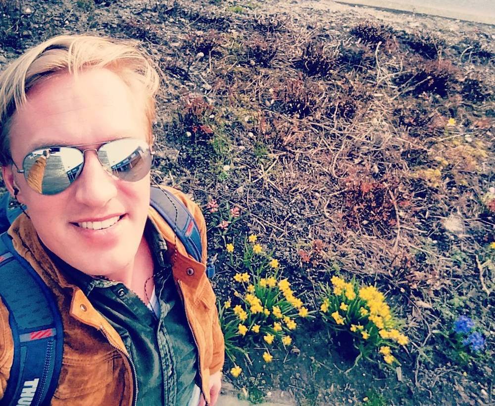 Selfie of a man in sunglasses and an orange jacket kneeling by yellow spring flowers on a slope.