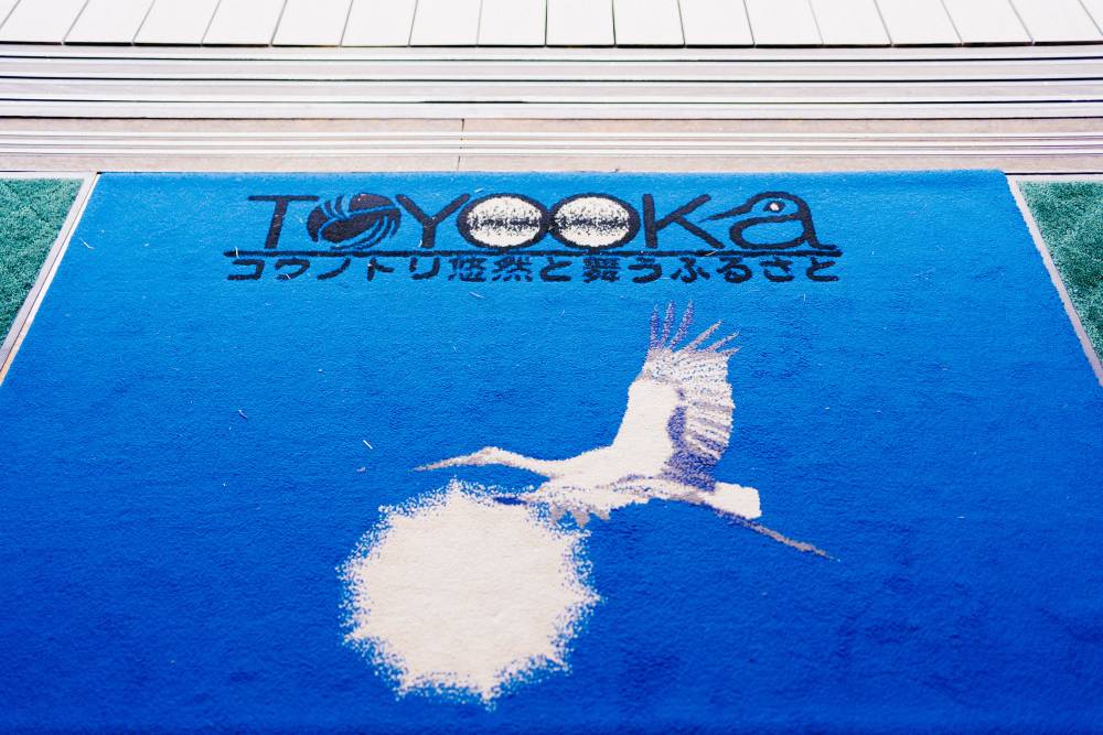 A floor mat for the Toyooka stork preserve. It’s a vibrant blue, with a stork on it, and the name of the city Toyooka in black.