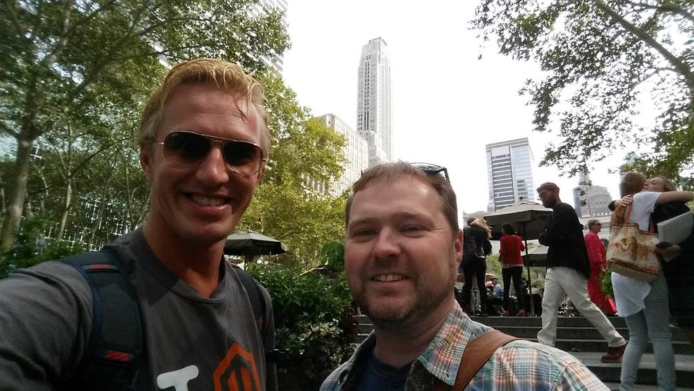 Two friends smile for a photo in Bryant Park with Midtown towers behind them.