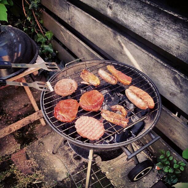 Burgers and sausages grilling on a charcoal barbecue in a garden.