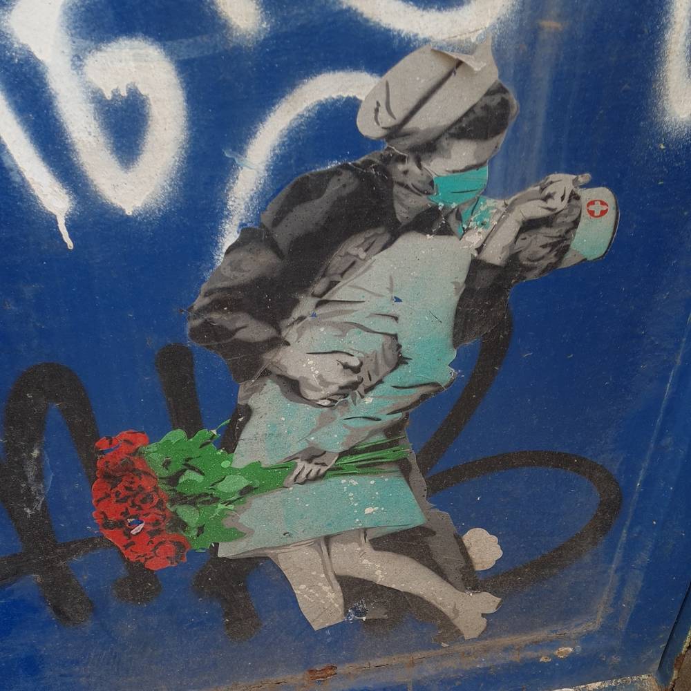 A sailor and a nurse, both wearing medical protection mask, trying kissing each other. With the nurse holding a bunch of flowers.