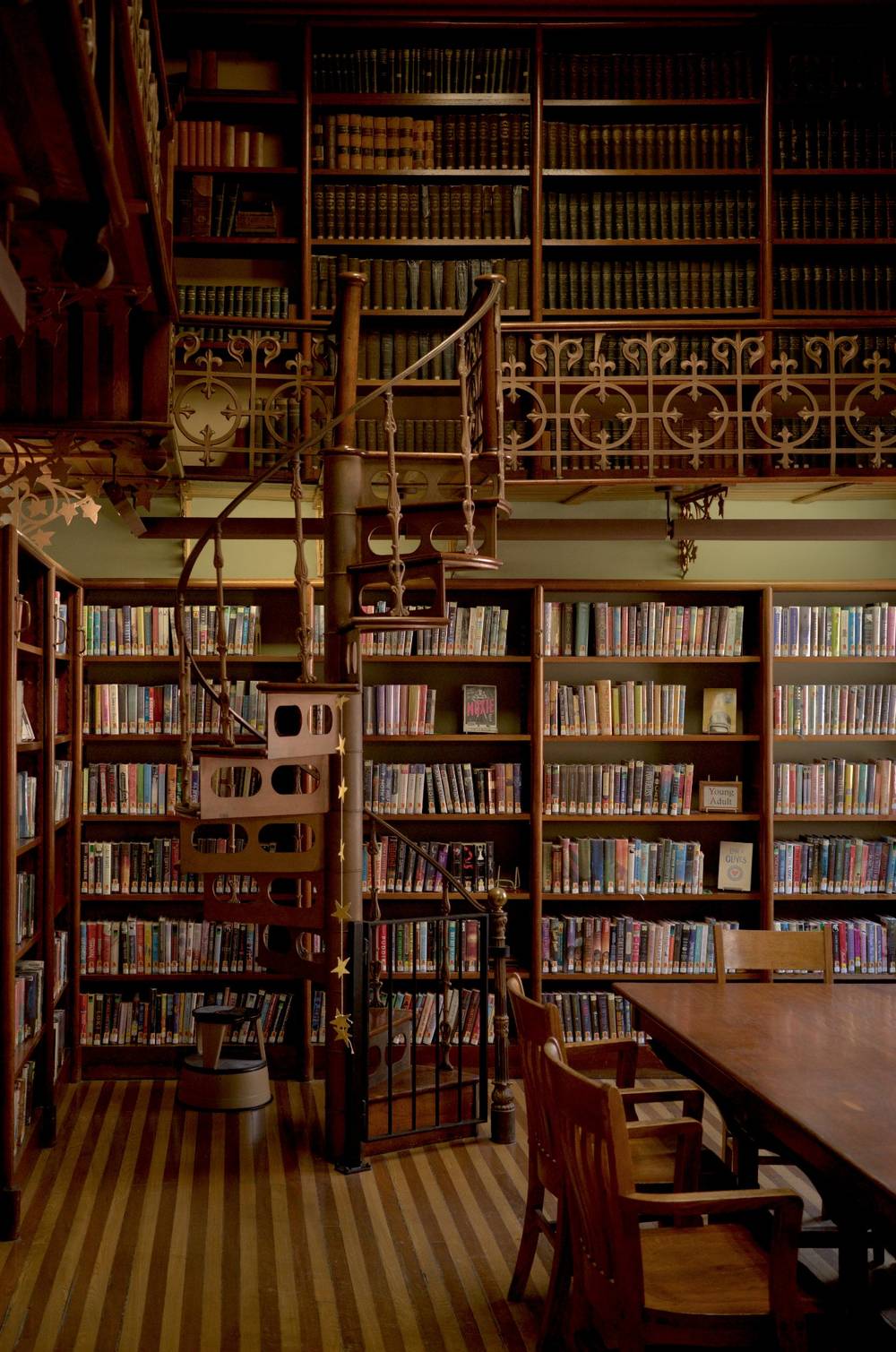 A library room with alternating cherry and oak flooring slats, a large wooden table with wooden chairs, floor to ceiling bookshelves with a spiral staircase leading to a second level of bookshelves.