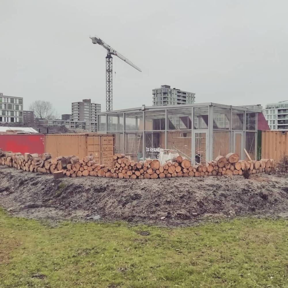 A construction site with a glass structure being built, logs stacked nearby.