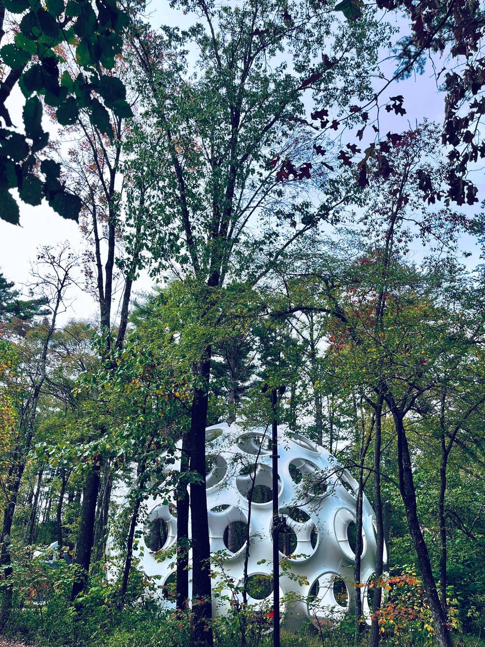 A large, white spherical sculpture with circular cutouts sits nestled among trees. The structure is partially obscured by green and early autumn foliage, under an overcast sky.