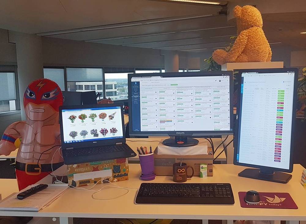 An office desk with a laptop, two monitors, a teddy bear on top, and a wrestler figurine.
