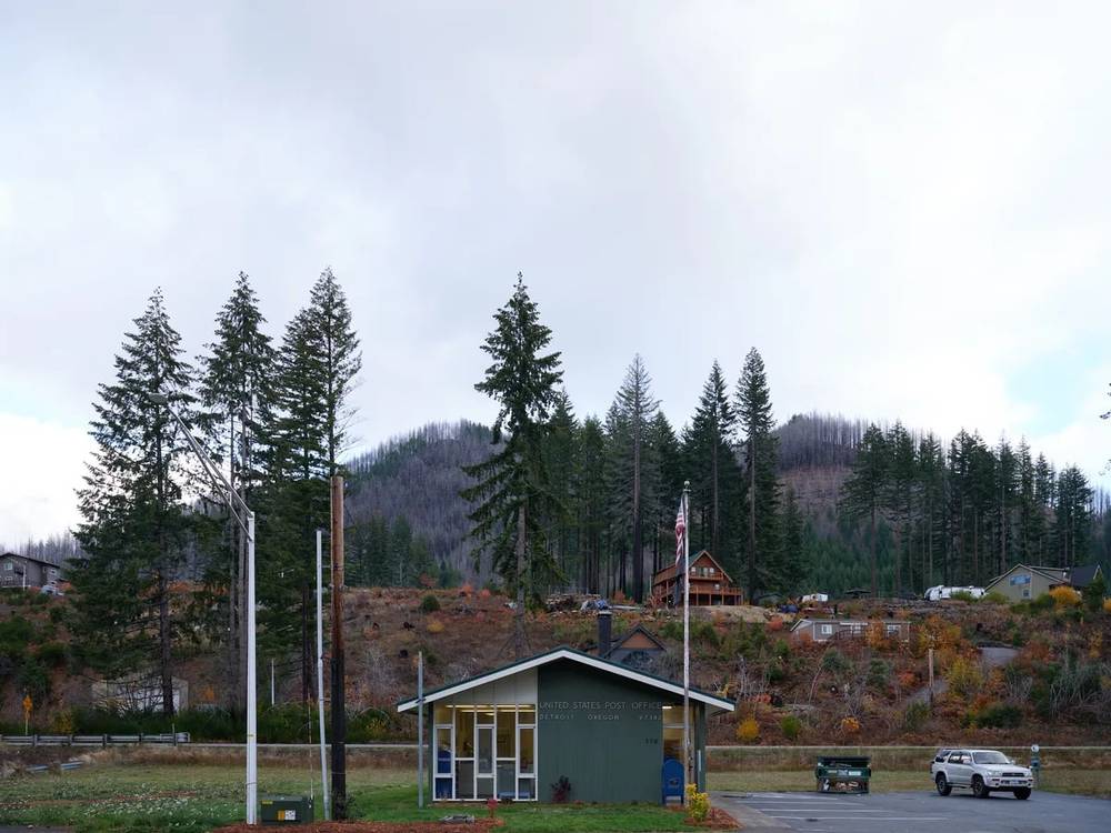 A modest single-story United States Post Office sits beneath towering evergreen-covered hills in Detroit, Oregon, its sage green exterior echoing the forest's palette. The building stands as a quiet sentinel of rural connectivity, with an American flag marking its federal presence against the dramatic backdrop of the Cascade foothills. Autumn's touch is visible in the scattered deciduous trees dotting the hillside, their amber and rust tones creating a warm counterpoint to the dominant conifers. The overcast sky and utility poles frame this quintessential small-town scene, where government service meets wilderness.