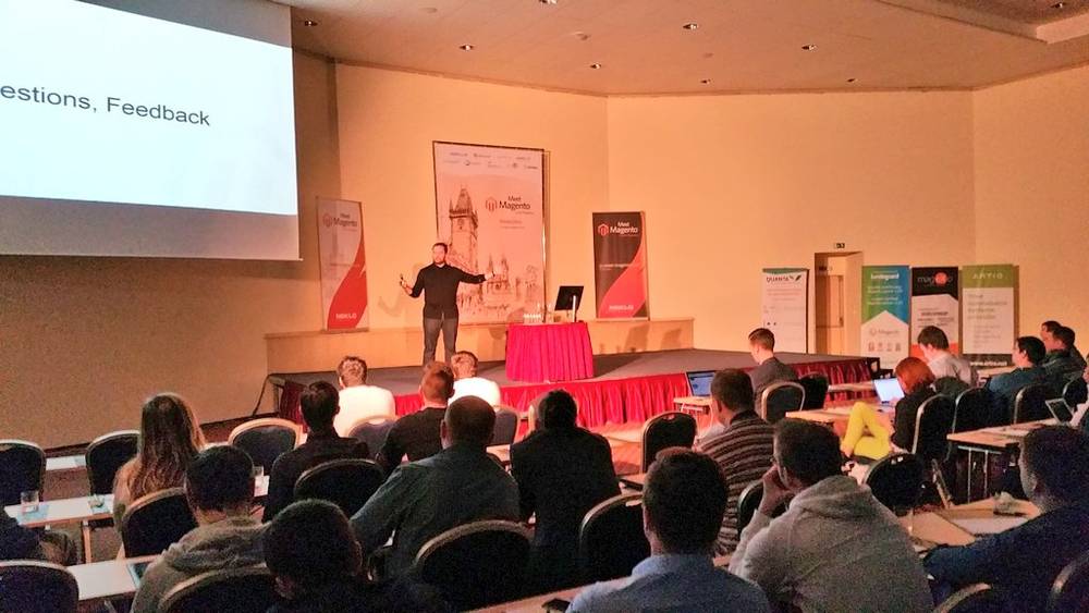 A speaker addresses a seated audience from a stage with Meet Magento banners.