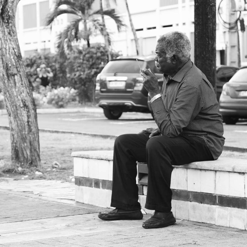 Black and white photo of an older man sitting on a bench in the street, smoking.