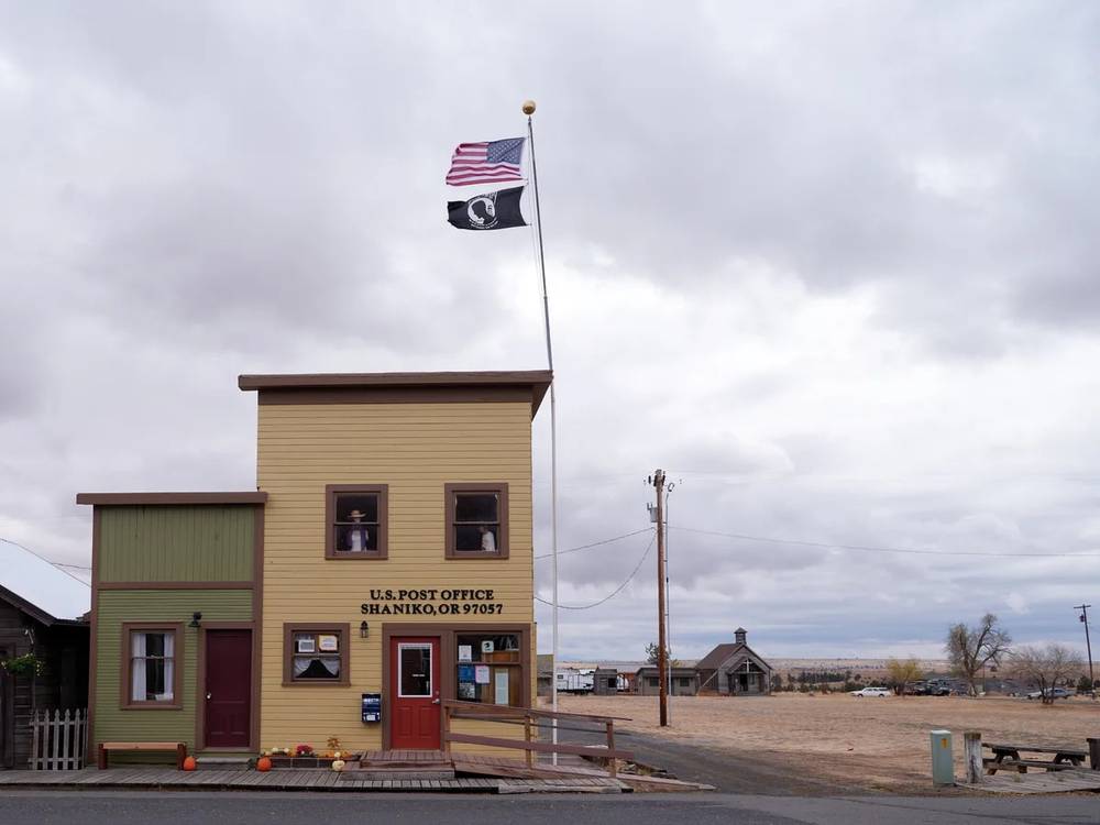 The weathered yellow clapboard post office of Shaniko, Oregon stands resolute beneath a blanket of pewter clouds, its American and POW-MIA flags snapping in the high desert wind. The modest frontier building, with its sage green trim and red door, anchors a desolate stretch of the Shaniko Historic District where time seems suspended between past and present. Autumn pumpkins dot the wooden boardwalk while telephone poles stretch into the vast Oregon plateau, their lines cutting through the moody overcast sky that threatens rain over this ghost town remnant.