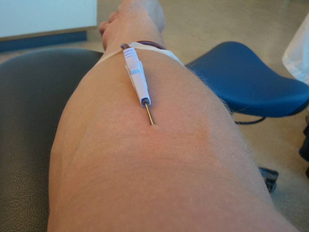 A close-up shows a needle taped to an arm during a blood donation.