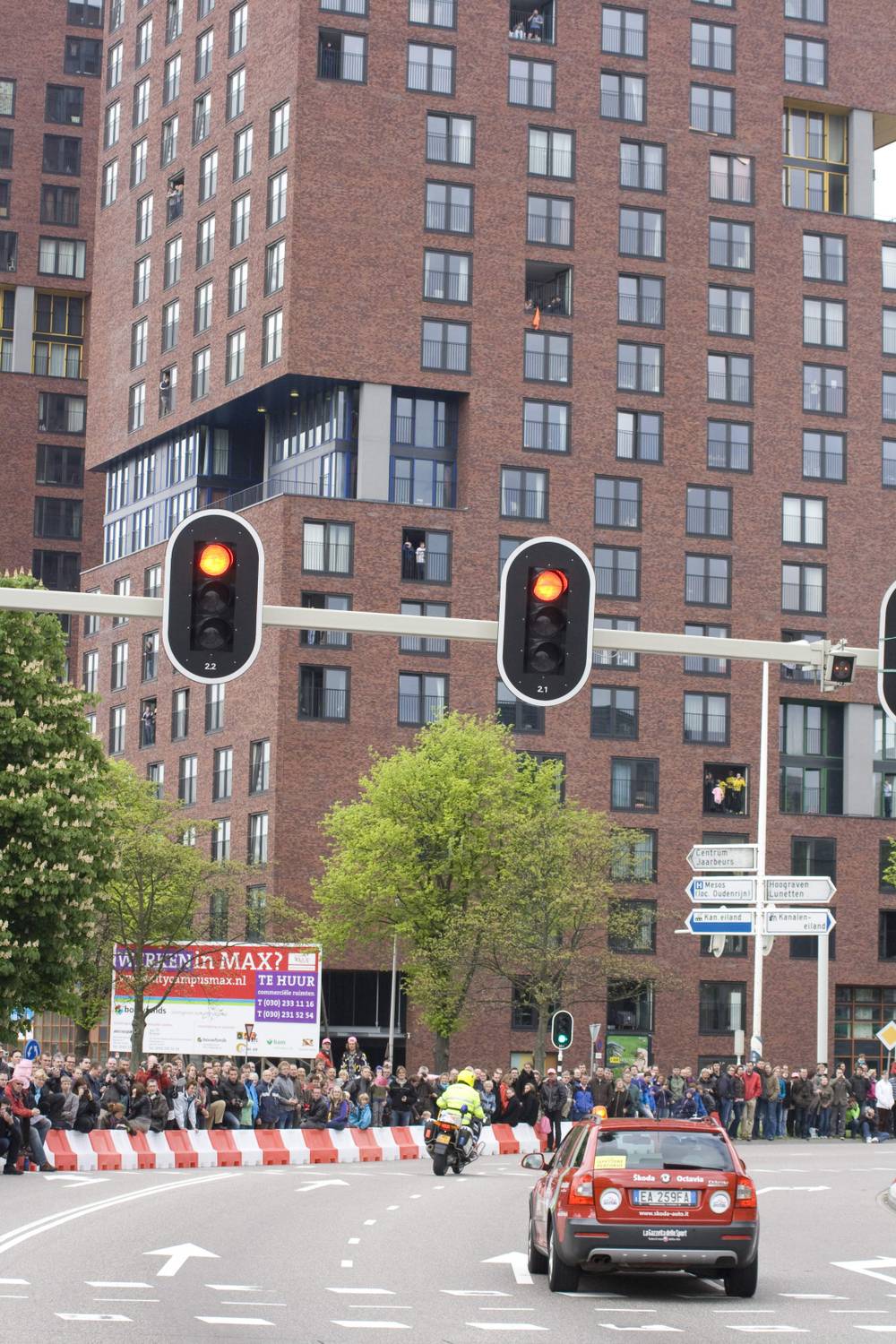 A crowd lines a street below a tall apartment block as a race car passes under traffic lights.