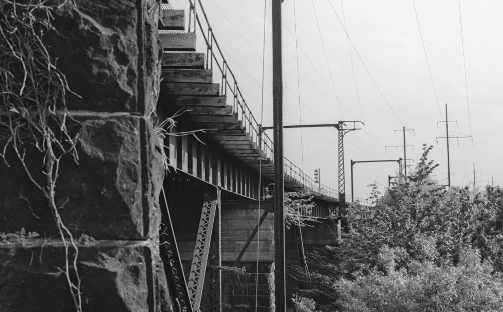 View along the underside of an electrified rail bridge from beside its stone abutment. Rough-cut stone, draped with dry vines, rises at left. A riveted steel plate girder carries wooden ties along the deck above. Beyond the bridge, catenary masts and tall steel transmission towers march into the distance, strung with overhead wires against a pale sky. Brush and small trees fill the lower right.