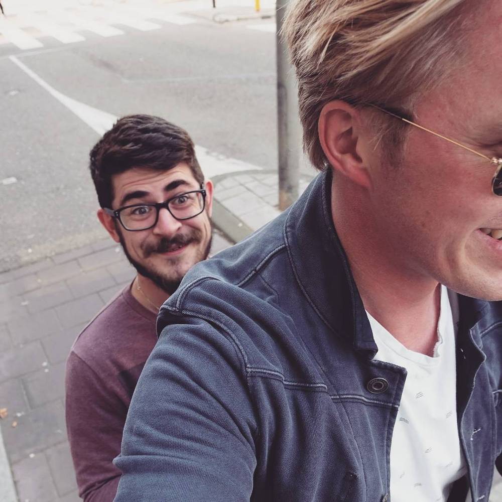Two men, one with glasses and a moustache, standing together on a street.