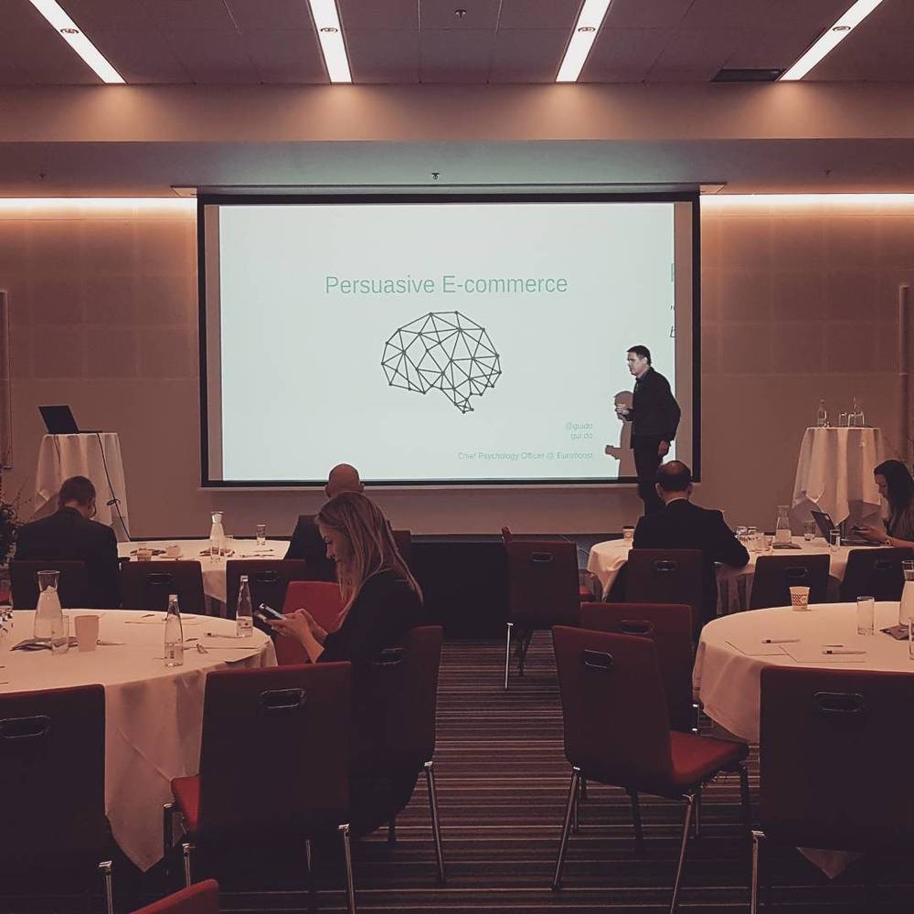 A conference presentation titled "Persuasive E-commerce" being given to seated attendees.