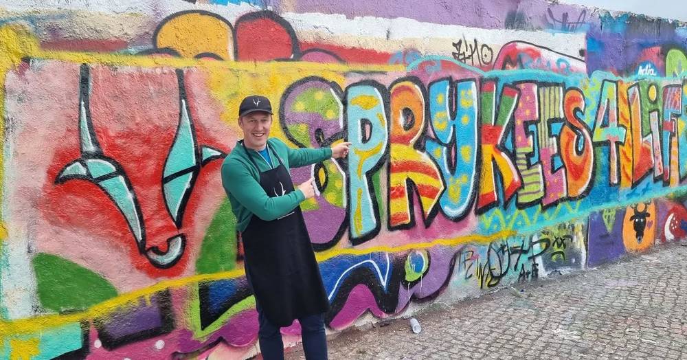 A man in a green sweater and apron pointing at a colorful graffiti mural reading "Spryketes4life" with a goat logo.