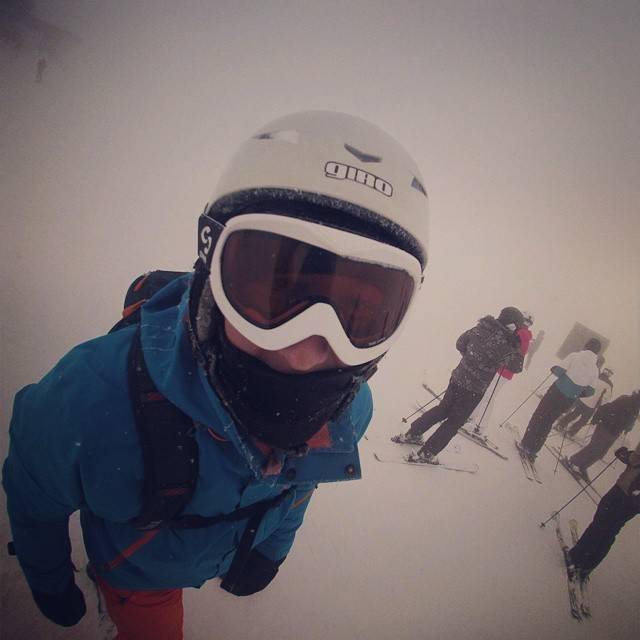 A person in ski goggles and a blue jacket photographed from above, with other skiers nearby.