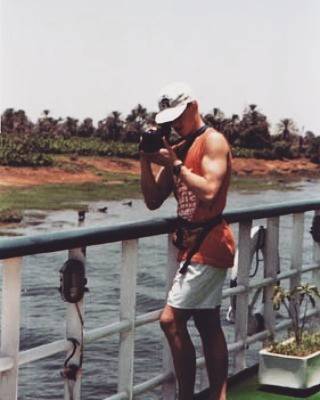 An old photo of a shirtless teenage boy taking a photo from a boat railing on a tropical river.