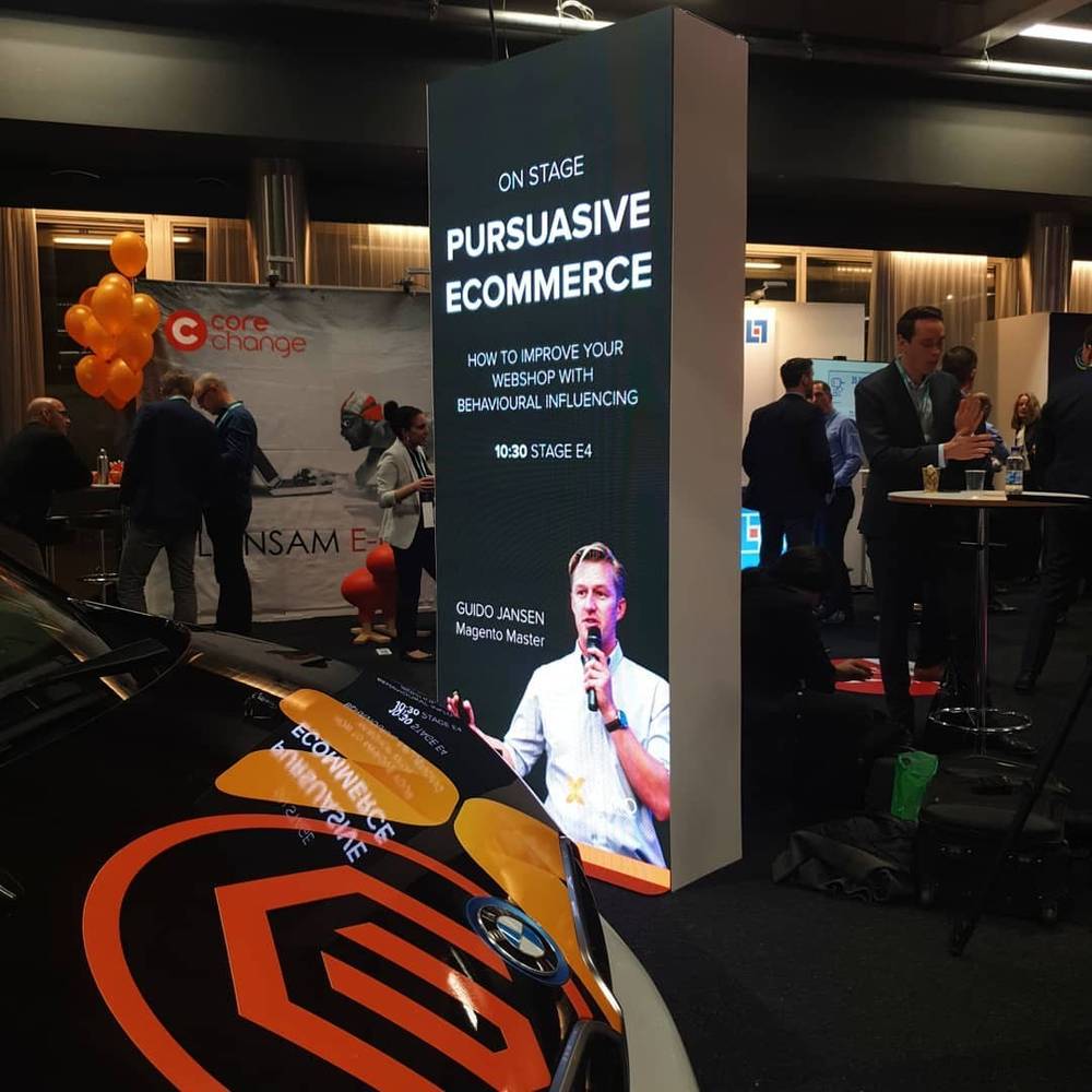 A man presents on stage titled "Persuasive Ecommerce" at a conference booth under illuminated signage.