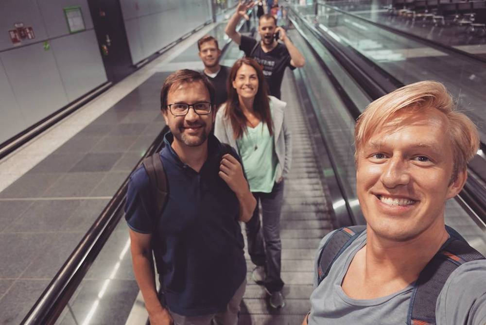 A group of colleagues on an airport moving walkway, one waving, taking a selfie.