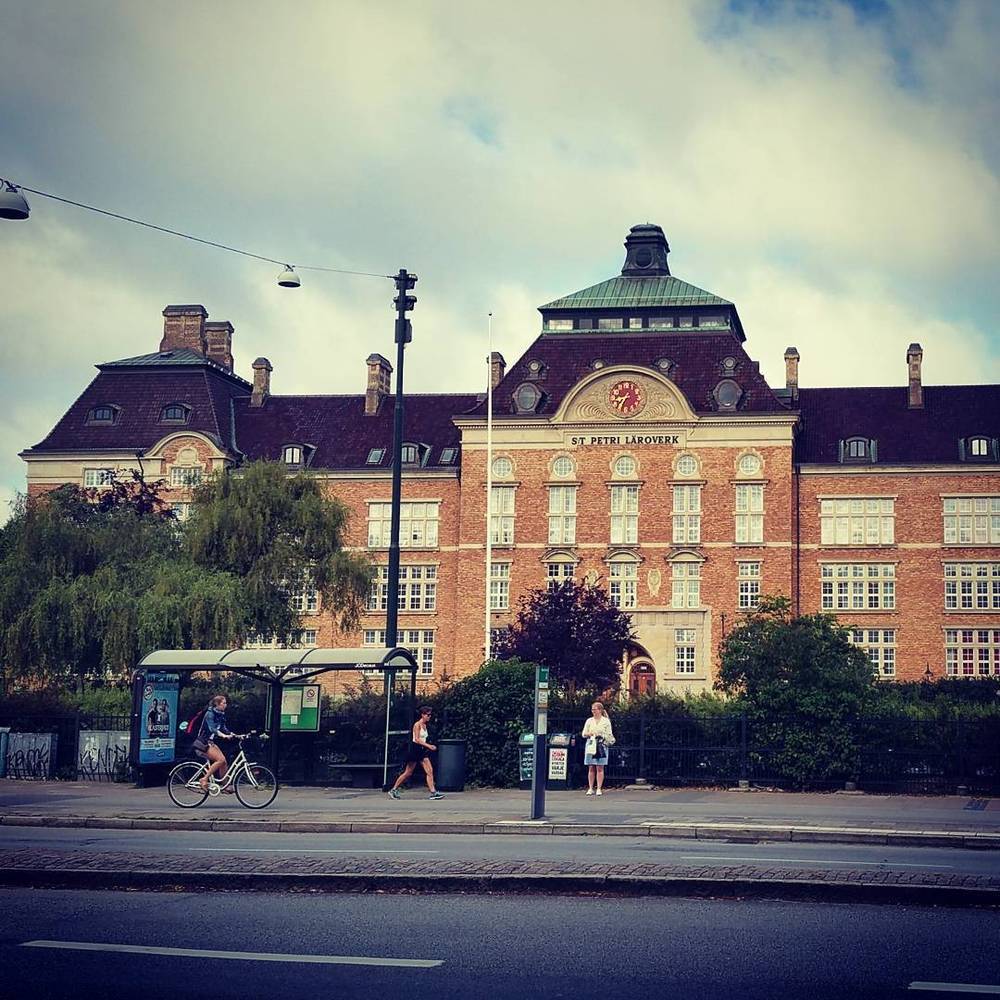 A red-brick historic school building with a clock tower, a bus stop and cyclist in front.